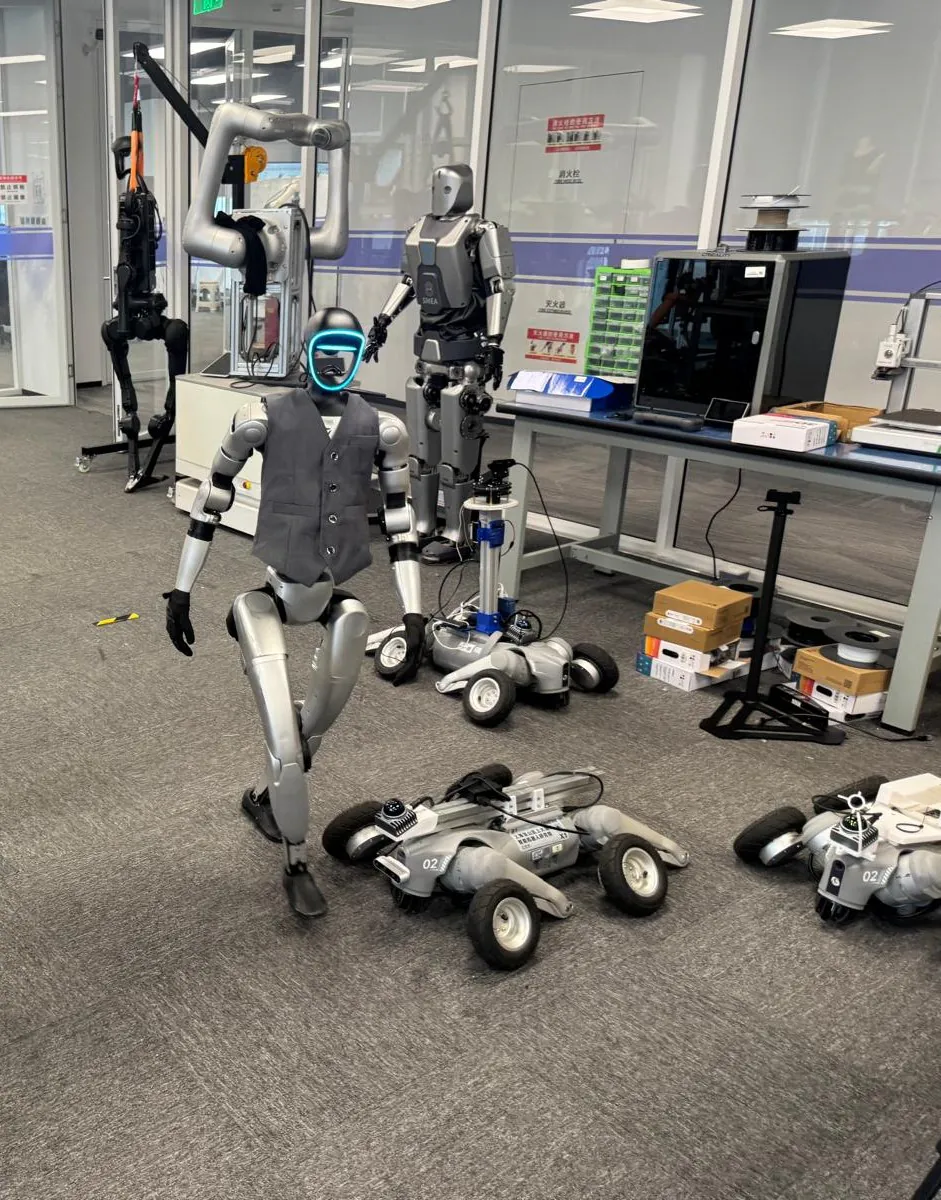 A Unitree humanoid in a gray vest stands among wheeled robots, with a second humanoid and a legged platform behind it in the lab